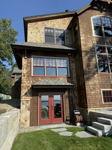 Multi-story shingle-style home with dark, weathered wood siding and red-trimmed windows, mid pressure-washing with hoses running across the lawn and patio.