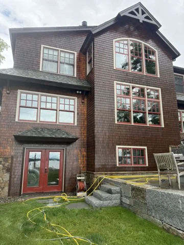 Same shingle-style home after staining, showing warm, even golden-brown wood siding with red trim, red French doors, and a finished stone retaining wall and patio.